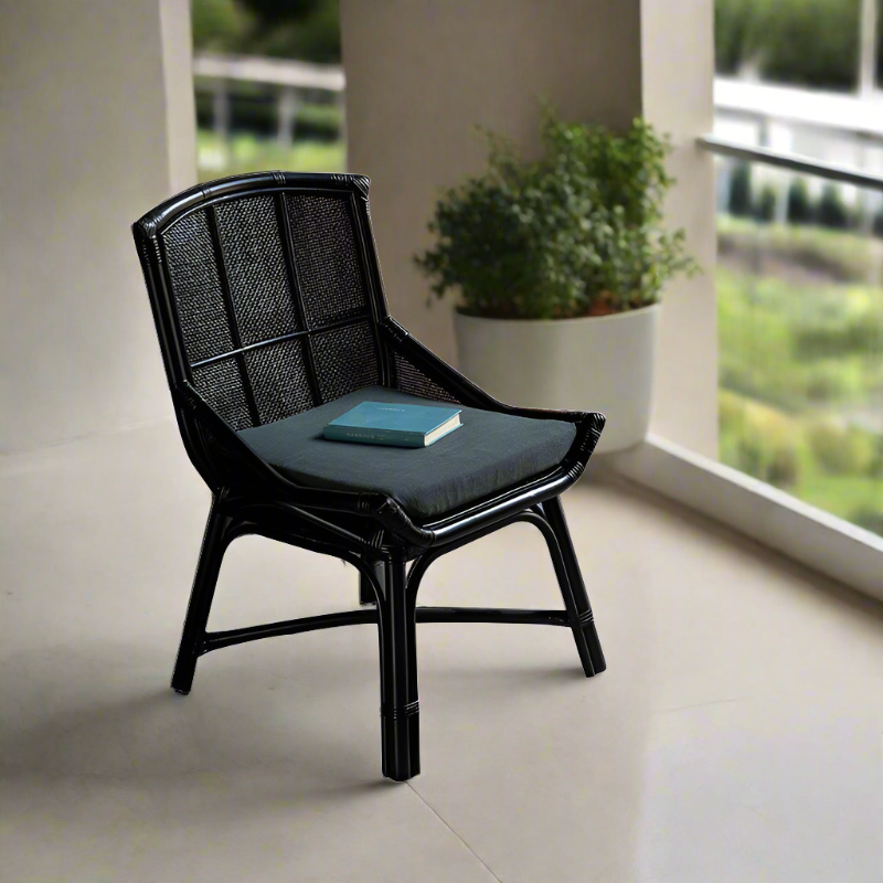 Black rattan dining chair with a flat seat and backrest, book on the seat, placed in a modern room with a balcony and plants in the background.