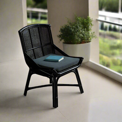 Black rattan dining chair with a flat seat and backrest, book on the seat, placed in a modern room with a balcony and plants in the background.