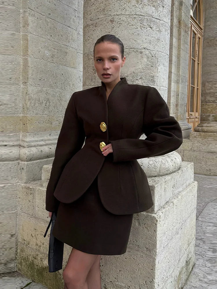 Outdoor front view of a deep brown tailored jacket and mini skirt set, styled with a sleek bag beside a stone building.