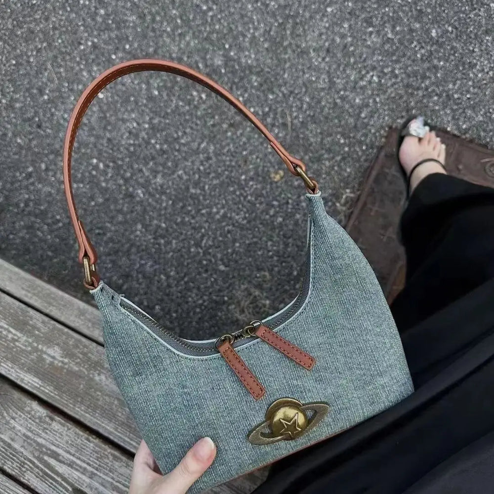 Blue denim handbag with brown leather strap held by a person on a textured surface.