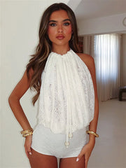 A woman wearing a white lace halter playsuit with an open back and grey shorts indoors.