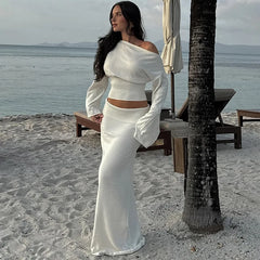 Woman in a white one shoulder flare sleeve sweater knit skirt set styled for a beach holiday standing on a beach with ocean and palm tree in the background