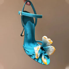 Women’s buckle strap formal summer heel in blue with a unique toe accent with a decorative flower on a beige background.
