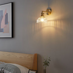 A wall-mounted LED lamp with a gold finish and a transparent glass shade, hanging over the bed.