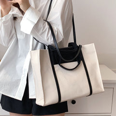 A canvas crossbody tote bag with black leather handles and a tan patch on the front, displayed against a neutral background, carried by a model in her hand.
