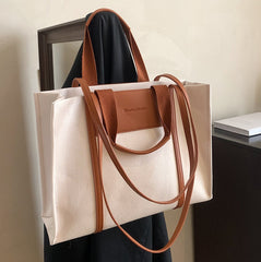 A canvas crossbody tote bag with brown leather handles and a tan patch on the front, displayed against a neutral background.