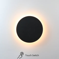 A circular acrylic LED wall-mounted light with a touch switch, illuminated and casting a warm white light on a wall surface with touch switch.