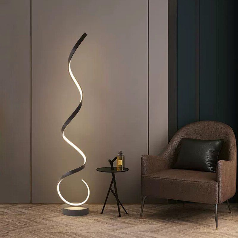A modern black floating LED floor lamp with an illuminated golden spiral design, placed in a room with a grey wall and a  chair and small round side table  next to it.