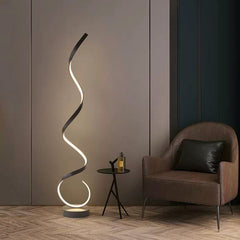A modern black floating LED floor lamp with an illuminated golden spiral design, placed in a room with a grey wall and a  chair and small round side table  next to it.