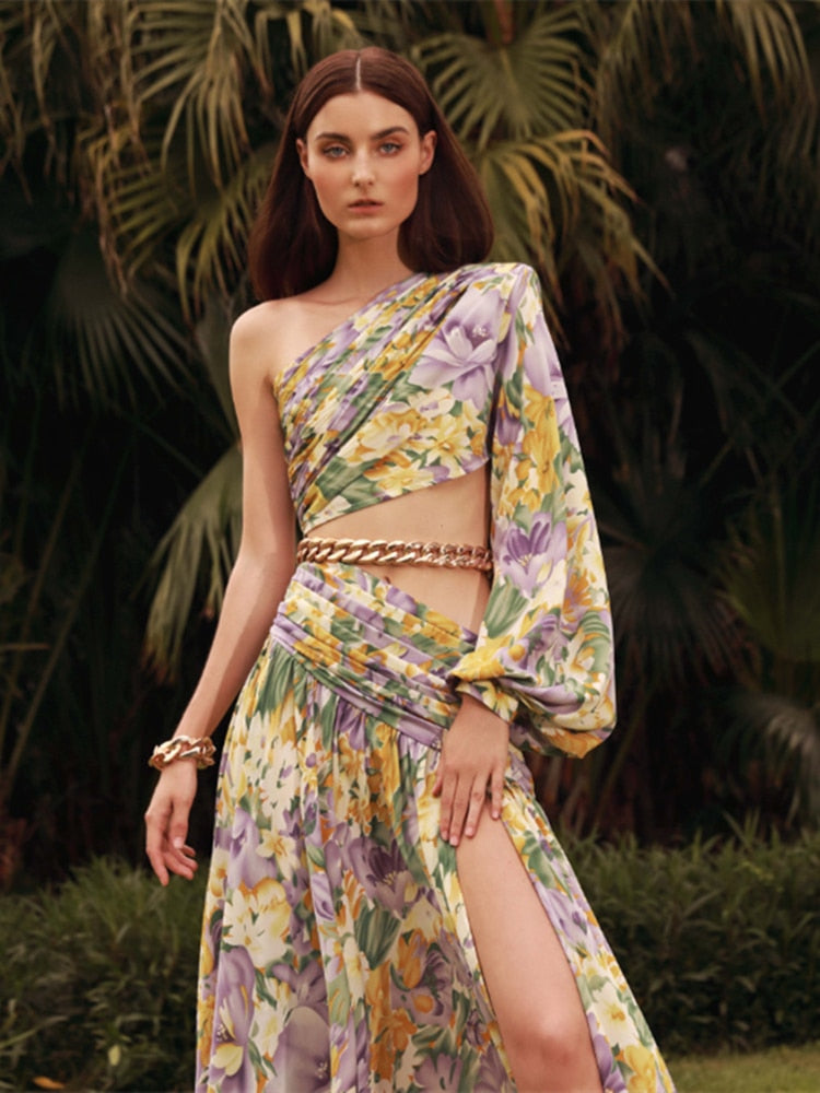 A model wearing a floral printed one-shoulder chain maxi dress with gold-tone chain detail and a side hollow detail.