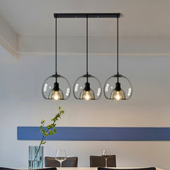 Three pendant ceiling lights with a black frame and clear glass shades hang over a white dining table with a plant and glasses on it, in a room with blue walls.