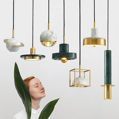 A collection of modern pendant lights with marble and copper finishes, some with a gold color, suspended from the ceiling. A woman with red hair is standing in the foreground with a large green leaf in her hand.